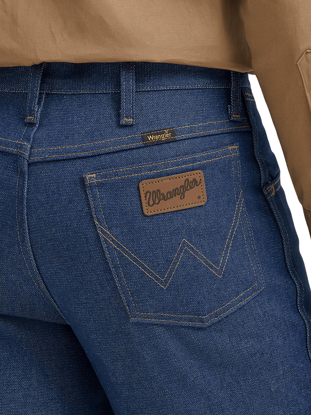 Close-up of the back waistband and pocket area of blue denim jeans, showing a brown leather Wrangler patch on the back pocket and a small yellow Wrangler tab near the waistband, with tan fabric at the top.