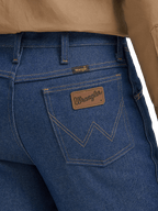 Close-up of the back waistband and pocket area of blue denim jeans, showing a brown leather Wrangler patch on the back pocket and a small yellow Wrangler tab near the waistband, with tan fabric at the top.