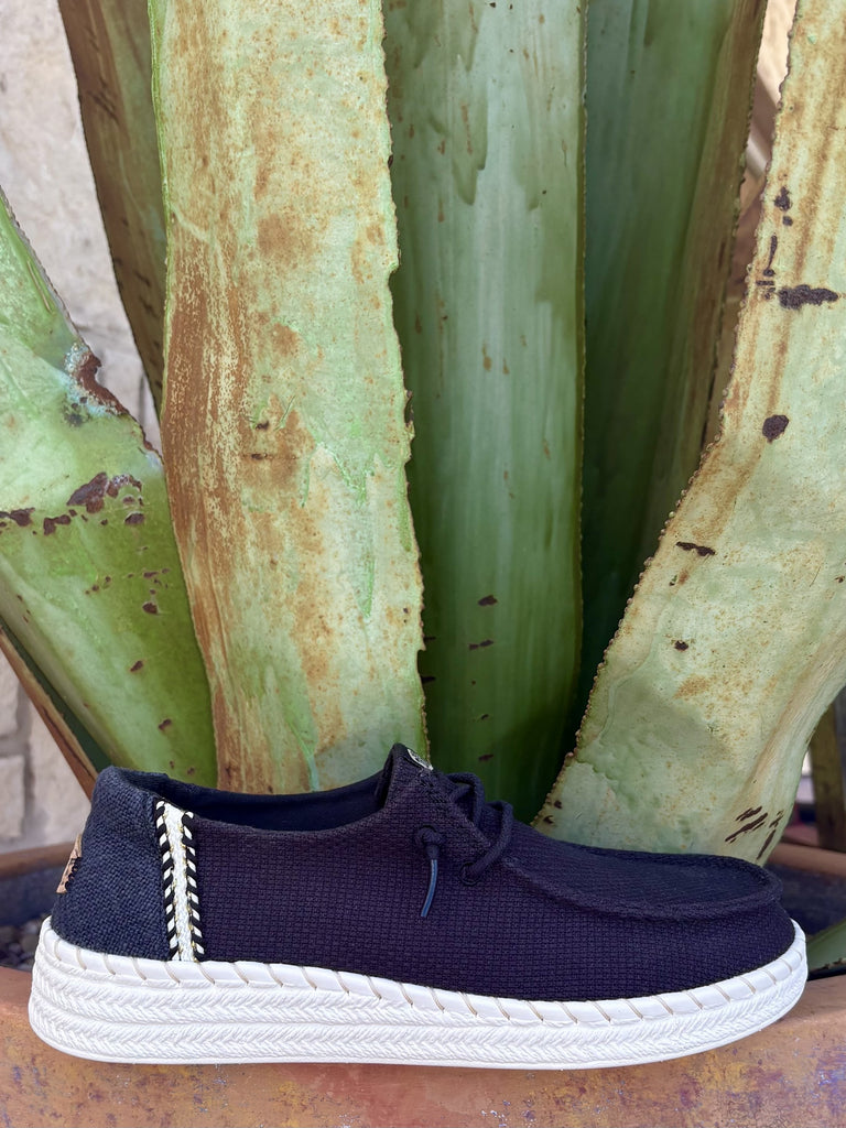 Women's black textured stretch canvas wide-fit moc-toe slip-on with black toggle lace, black-and-white whipstitch side seam detail, brown leather Hey Dude oval logo patch, and chunky white braided-texture EVA outsole — Blair's Western Wear Marble Falls TX