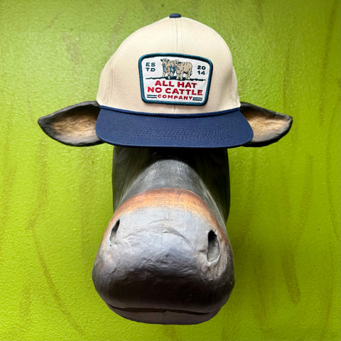 All Hat No Cattle Company cream and navy cap, structured 6-panel snapback for adjustable fit, embroidered cattle patch with bold logo, breathable eyelet vents – ALL HAT CAP. Available at Blair's Western Wear, Marble Falls, TX.
