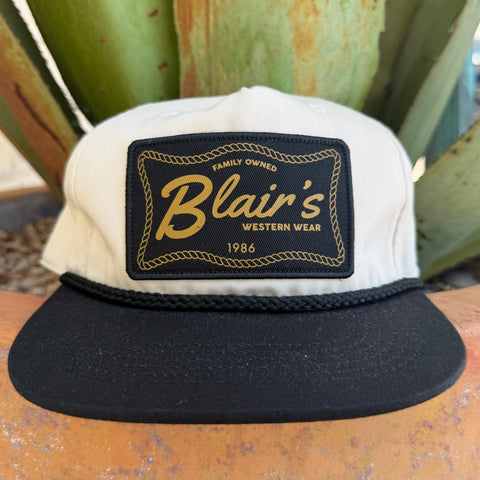 A black and off- white snapback cap with a prominent black front patch featuring gold embroidered text that reads 'Blair's Western Wear 1986'.