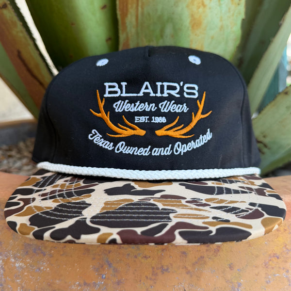 A black snapback cap with 'BLAIR'S Western Wear' embroidered on the front, featuring orange antler graphics and a brown, beige, and black camouflage patterned brim, set against a background of green succulent leaves.