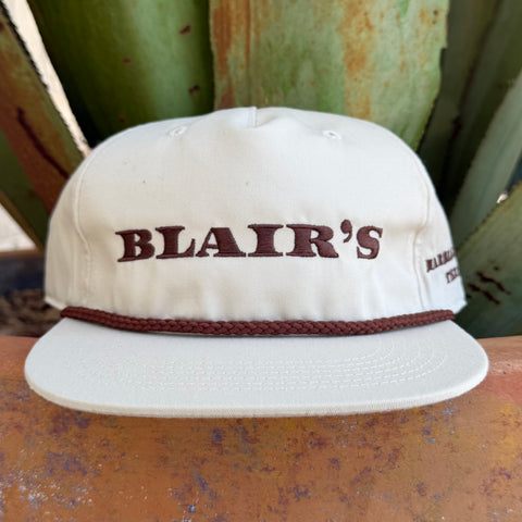 An off-white snapback cap with 'BLAIR'S' embroidered in dark brown across the front, featuring a matching brown braided rope detail around the crown and a flat brim