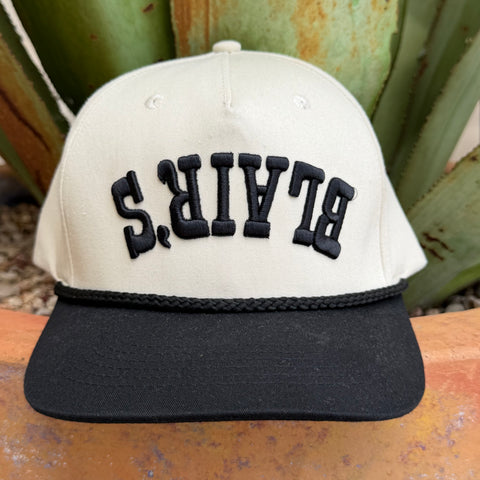 A structured baseball cap with an off-white crown and a black flat brim, featuring the word 'BLAIR'S' embroidered in black arched letters across the front updside down 