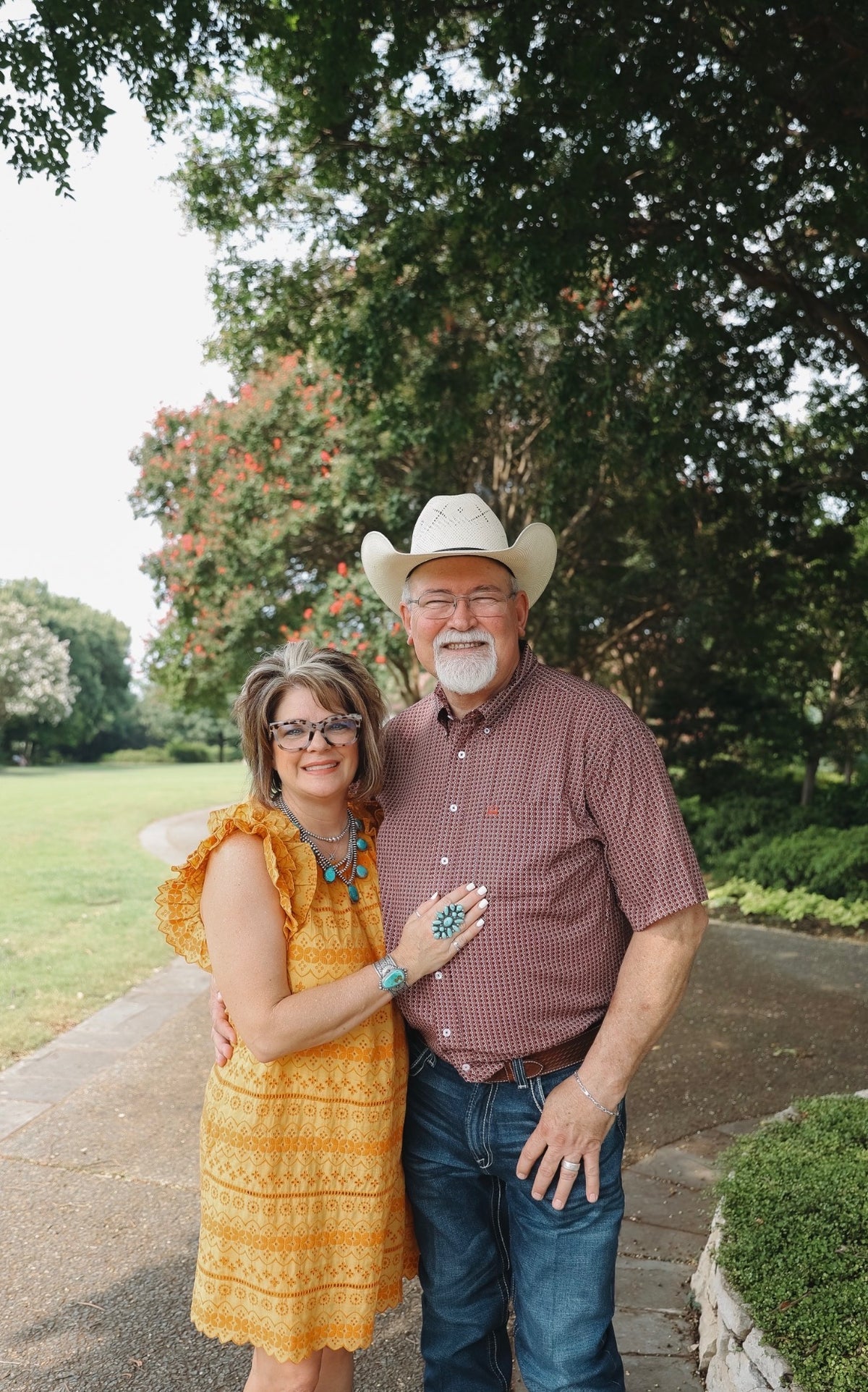 A couple standing outdoors on a paved path under a large tree. The woman wears a bright yellow patterned dress and turquoise jewelry, and the man wears a white cowboy hat, a red-and-white checkered button-up shirt, blue jeans, and a watch.