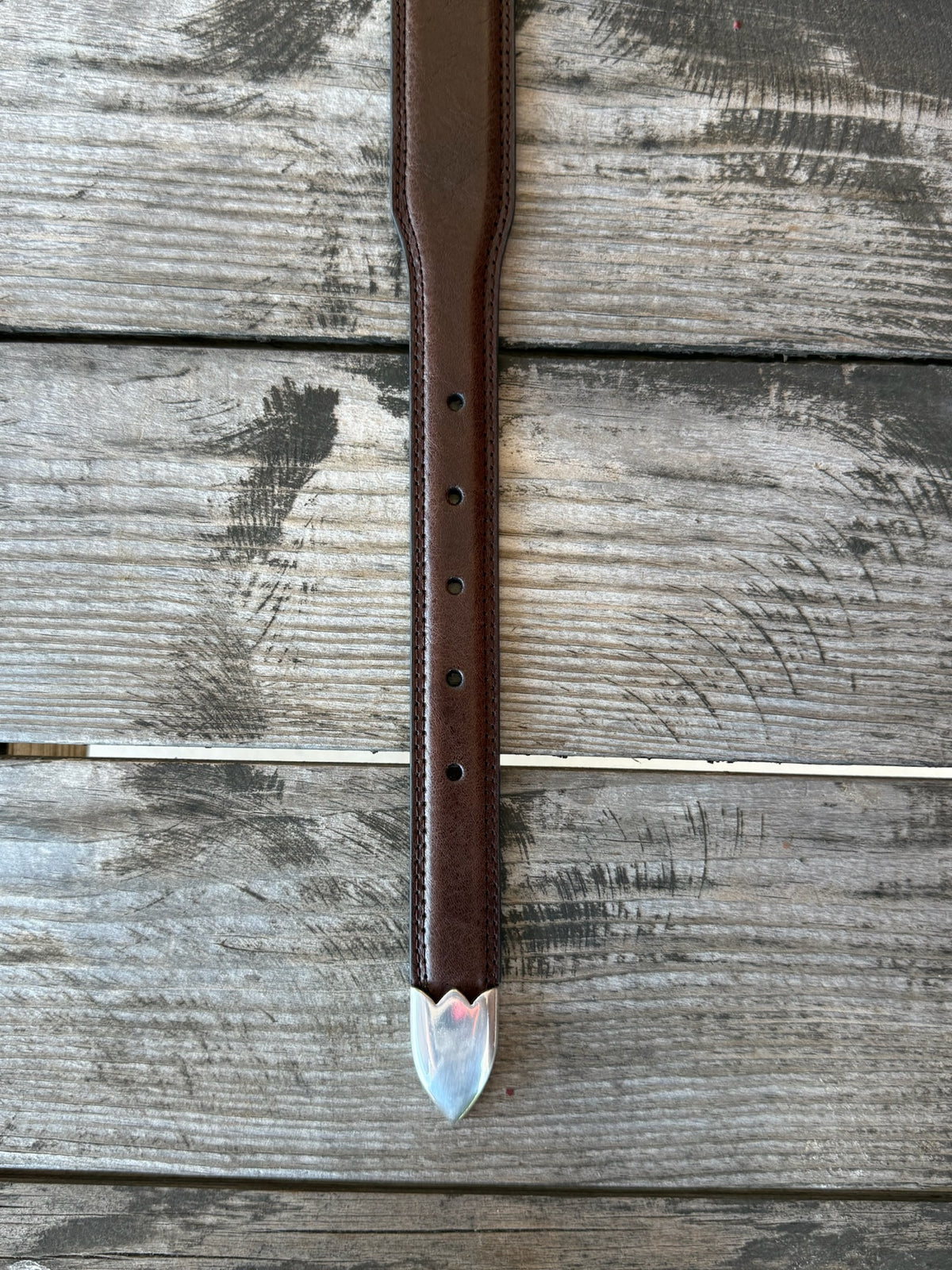 A brown leather belt with a silver-toned western-style buckle and decorative tip, coiled on a rustic wooden surface with a distressed red wooden background.