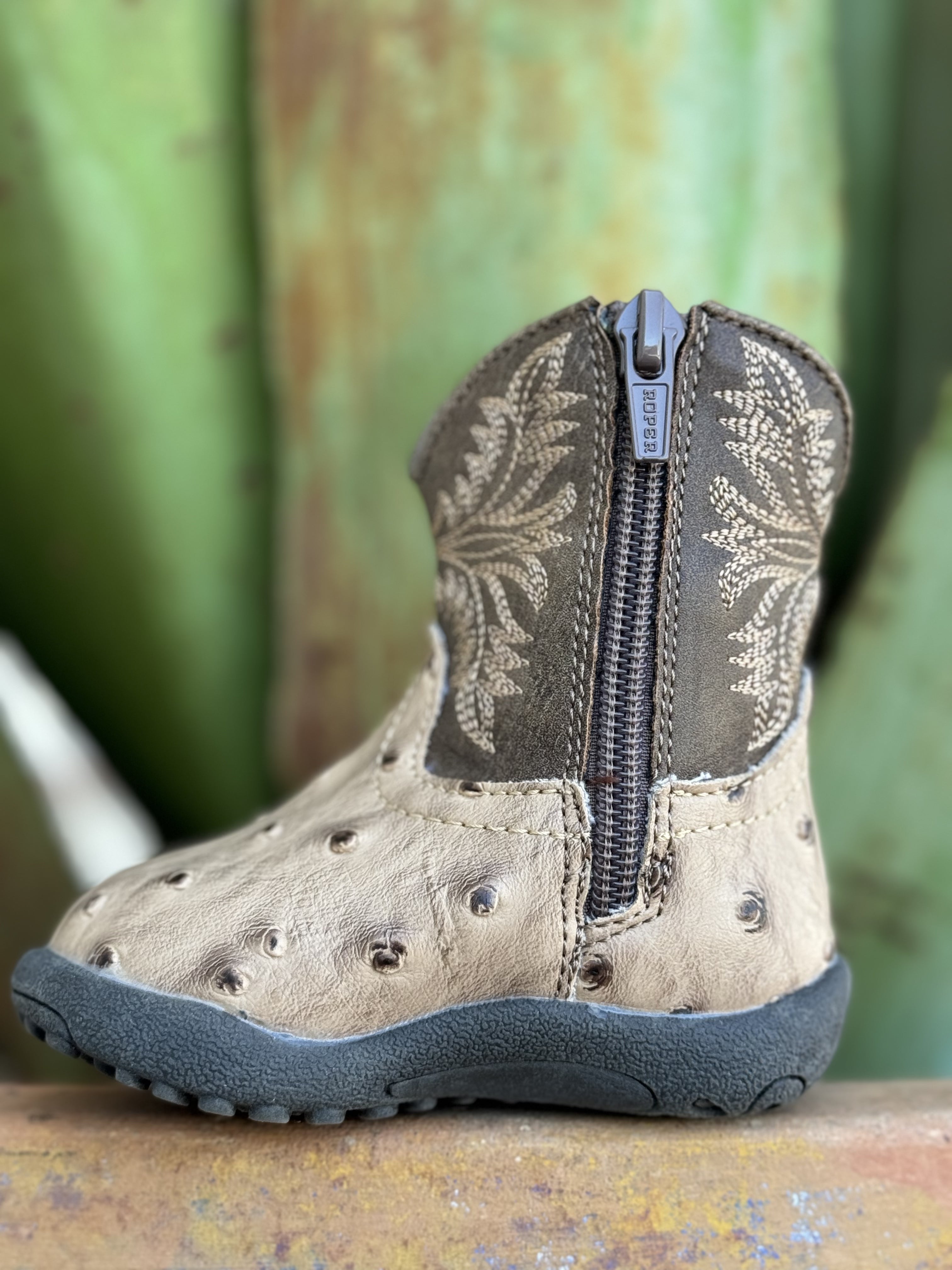 Introduce your baby to upscale Western wear with this charming Infant Baby Roper Boot! Made from leather with a stylish tan ostrich texture, this boot offers a luxurious look for tiny feet. Designed for ease and comfort, it features a handy side zipper for quick changes. The durable rubber sole provides excellent traction and flexibility for little explorers on the move.  Infant Roper Boot - 91612243500 - Available at BLAIR'S Western Wear located in Marble Falls TX 
