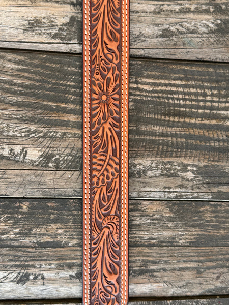 A tooled brown leather belt with a large engraved silver and gold buckle featuring the ARIAT logo, displayed on a rustic wooden surface with a weathered red wooden background. Only available at Bair's Western Wear located in Marble Falls, TX 