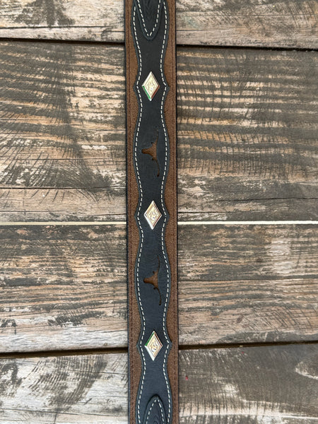 A western-style leather belt coiled on a rustic wooden surface, featuring a large engraved silver buckle with a steer design, a dark brown leather strap, and a black leather section with white stitching and a small diamond-shaped logo. It also features longhorn cutouts. Available at BALIR'S Western Wear located in Marble Falls TX 