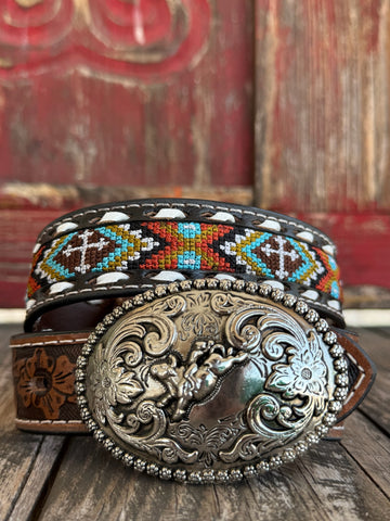 Kid's Aztec-Inspired Embroidered Cross Belt Black leather strap with a multicolor Aztec-inspired embroidered cross panel in teal, orange/red, olive/gold, and white. White lace whipstitch edge detail flanks the embroidered panel. Cognac floral scroll-tooled leather keeper loop. Large oval silver buckle with 3D bucking bronco and rider centerpiece, ornate floral scroll engraving, and beaded rope border. Available at Blair's Western Wear, Marble Falls, TX