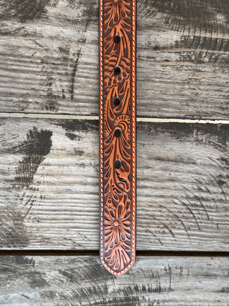 A tooled brown leather belt with a large engraved silver and gold buckle featuring the ARIAT logo, displayed on a rustic wooden surface with a weathered red wooden background. Only available at Bair's Western Wear located in Marble Falls, TX 
