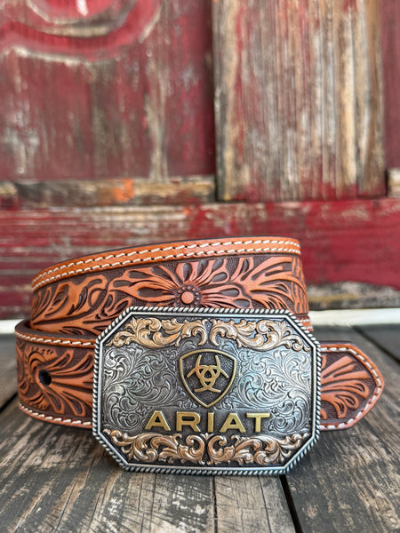 A tooled brown leather belt with a large engraved silver and gold buckle featuring the ARIAT logo, displayed on a rustic wooden surface with a weathered red wooden background. Only available at Bair's Western Wear located in Marble Falls, TX 