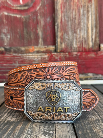 A tooled brown leather belt with a large engraved silver and gold buckle featuring the ARIAT logo, displayed on a rustic wooden surface with a weathered red wooden background. Only available at Bair's Western Wear located in Marble Falls, TX 