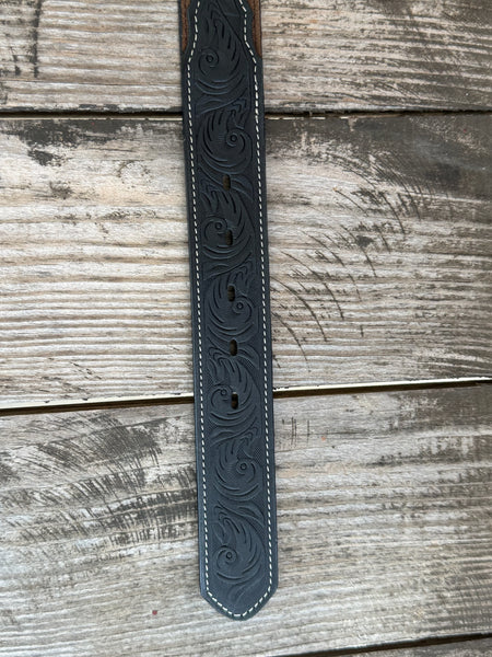 A western-style leather belt coiled on a rustic wooden surface, featuring a large engraved silver buckle with a steer design, a dark brown leather strap, and a black leather section with white stitching and a small diamond-shaped logo. It also features longhorn cutouts. Available at BALIR'S Western Wear located in Marble Falls TX 