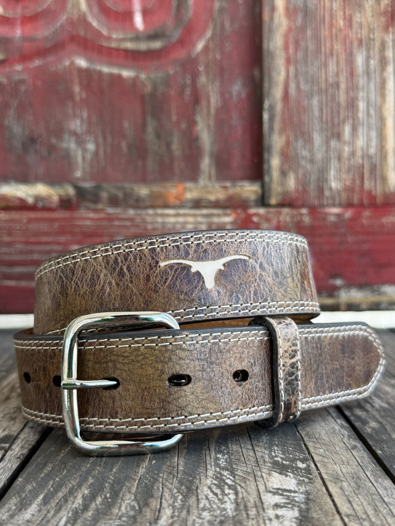 A brown distressed leather belt with a silver-toned buckle and a silver Longhorn logo, coiled on a rustic wooden surface with a weathered red wooden background. Available at Blair's Western Wear, Marble Falls, TX