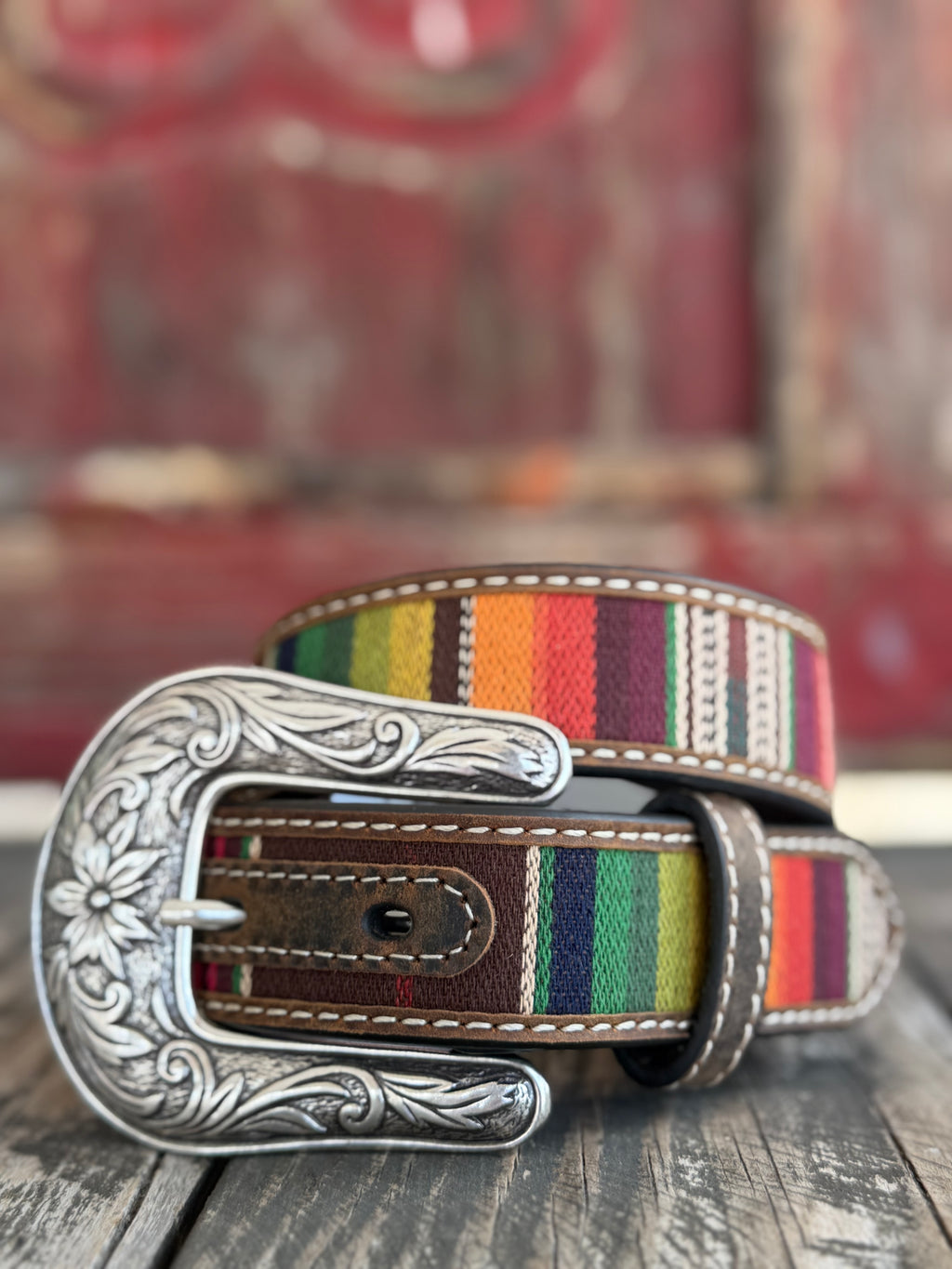 A vibrant multicolor serape stripe runs the length of this brown leather belt, edged with cream whipstitching and finished with a beautifully engraved floral silver buckle — bold southwestern color with classic western craftsmanship. Available at BLAIR'S Western Wear located in Marble Falls TX 