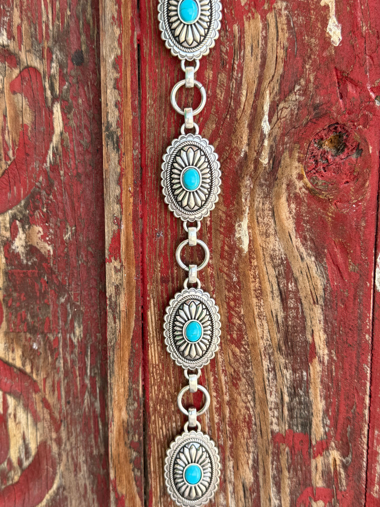 A1308233 — Kid's Turquoise Concho Chain Western Belt Full silver concho chain belt with repeating oval silver conchos featuring engraved petal/sunburst detail, rope edge border, and turquoise stone center in each. Silver O-ring links connect each concho. Adjustable silver O-ring chain tail with small turquoise stone drop tip. Available at Blair's Western Wear located in Marble Falls TX 