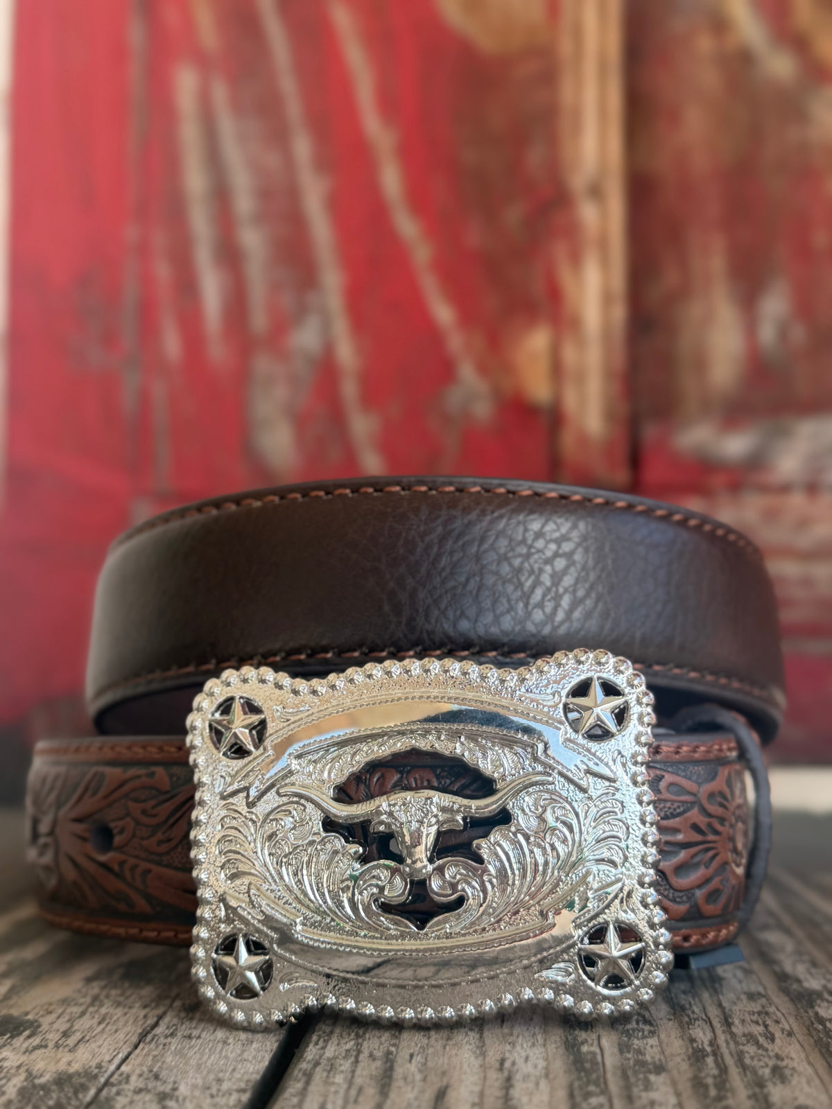 A dark brown leather belt with a highly detailed, engraved silver buckle featuring a western bull skull and star motifs, displayed on a rustic wooden surface with a red wooden background. Available at Blair's Western Wear located in Marble Falls TX