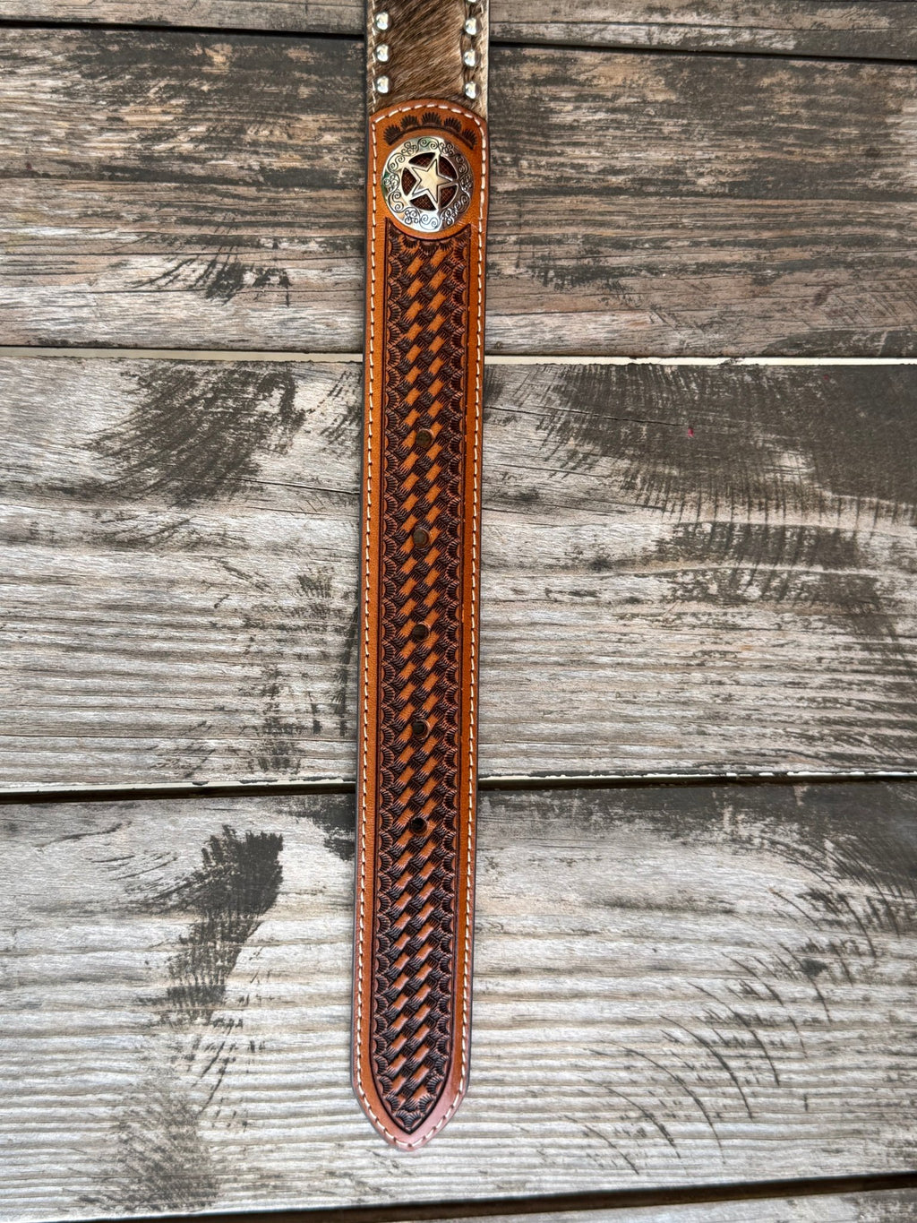 A long, narrow brown leather belt laid flat on a weathered gray wooden surface. The belt has a woven basketweave pattern in dark brown and black, a silver-toned concho with a star, and silver-toned metal rivets near the top.
