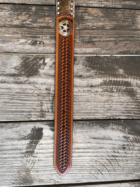 A long, narrow brown leather belt laid flat on a weathered gray wooden surface. The belt has a woven basketweave pattern in dark brown and black, a silver-toned concho with a star, and silver-toned metal rivets near the top.