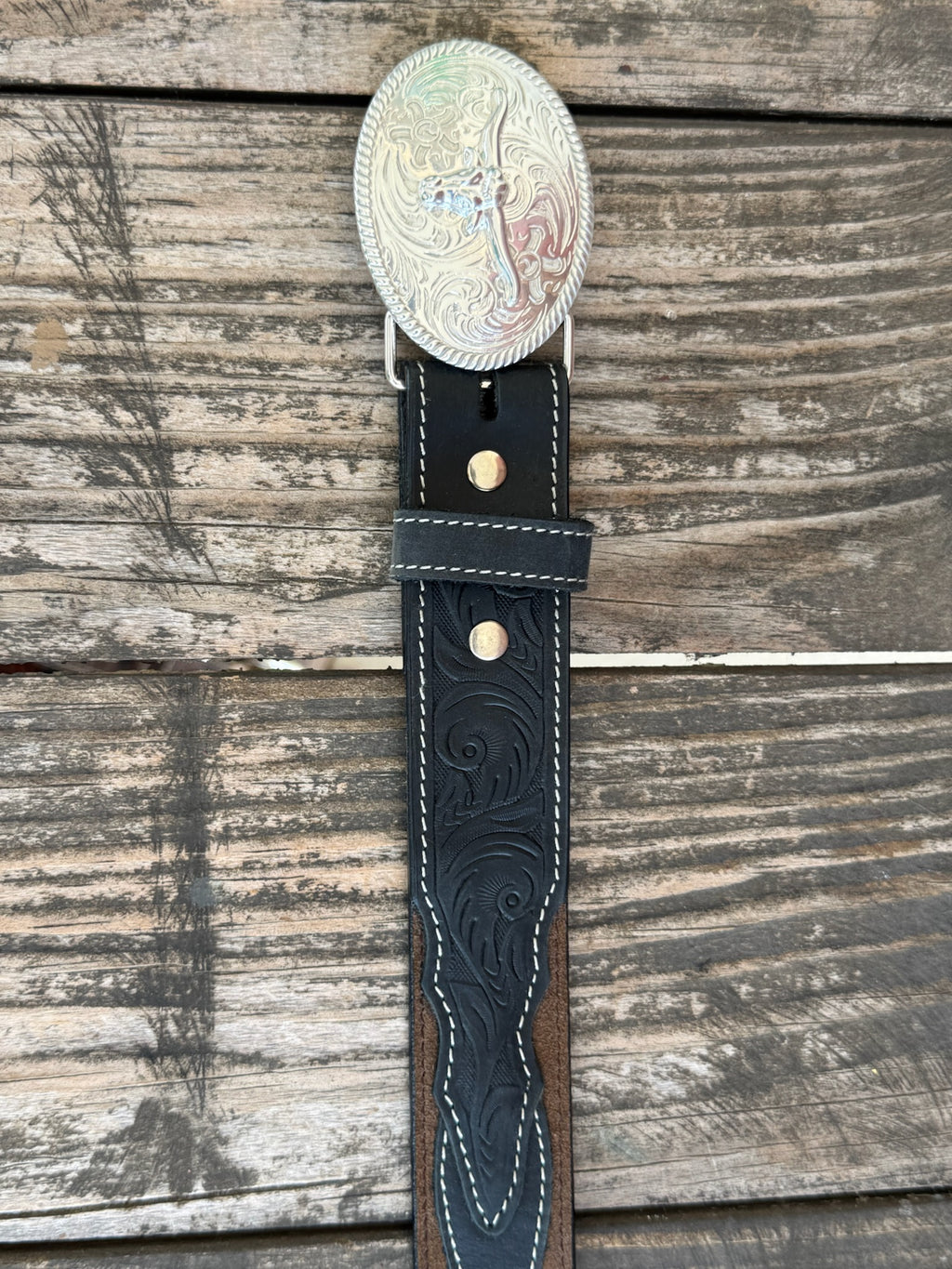A western-style leather belt coiled on a rustic wooden surface, featuring a large engraved silver buckle with a steer design, a dark brown leather strap, and a black leather section with white stitching and a small diamond-shaped logo. It also features longhorn cutouts. Available at BALIR'S Western Wear located in Marble Falls TX 