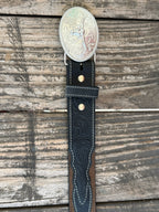 A western-style leather belt coiled on a rustic wooden surface, featuring a large engraved silver buckle with a steer design, a dark brown leather strap, and a black leather section with white stitching and a small diamond-shaped logo. It also features longhorn cutouts. Available at BALIR'S Western Wear located in Marble Falls TX 