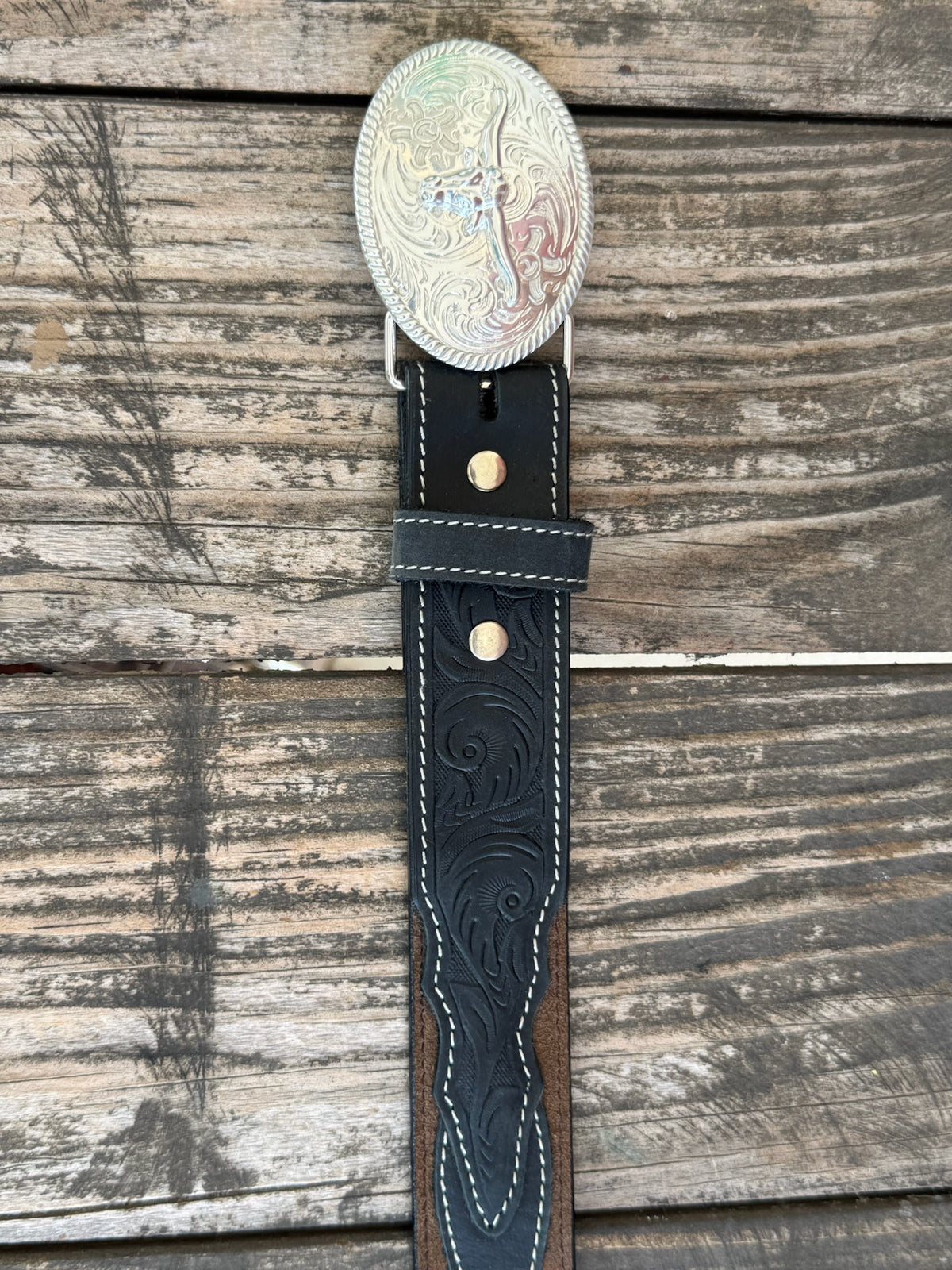 A western-style leather belt coiled on a rustic wooden surface, featuring a large engraved silver buckle with a steer design, a dark brown leather strap, and a black leather section with white stitching and a small diamond-shaped logo. It also features longhorn cutouts. Available at BALIR'S Western Wear located in Marble Falls TX 