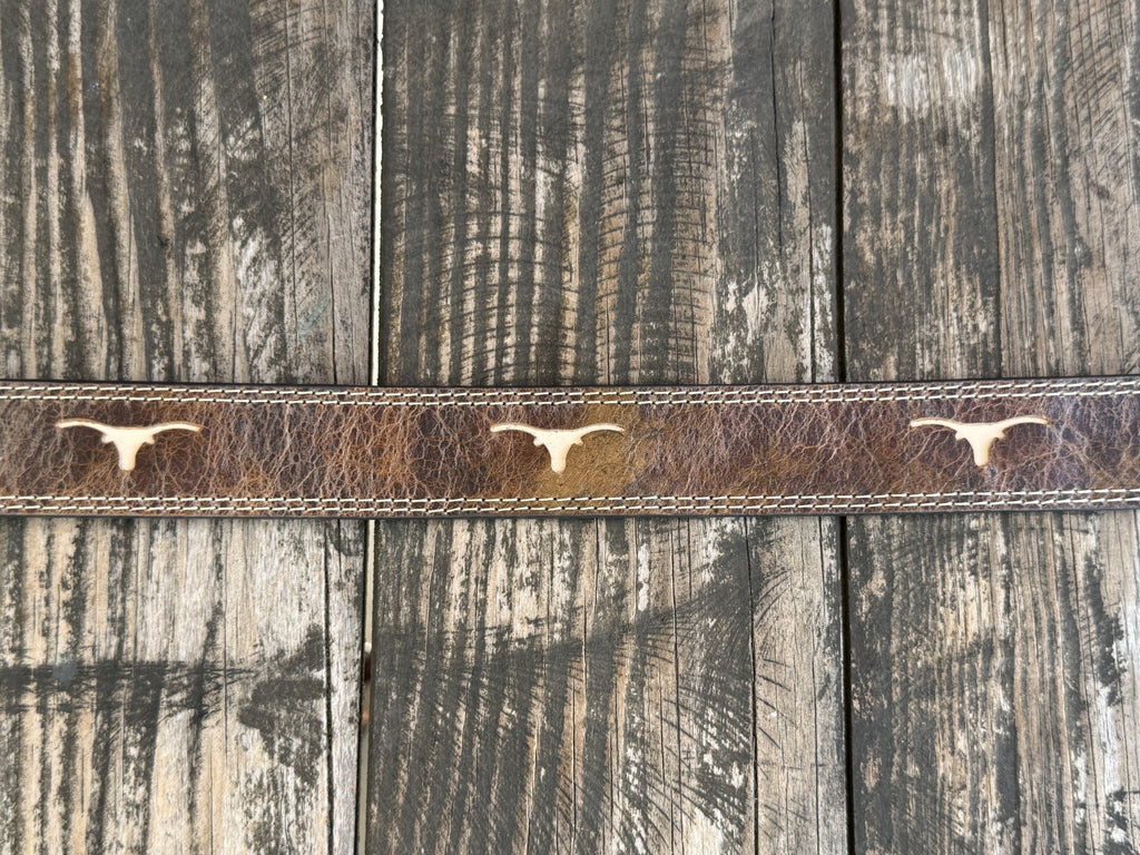 A close-up of a brown leather belt with three light-colored steer skull emblems, laid flat on a rustic weathered wood plank surface. Available at Blair's Western Wear, Marble Falls, TX