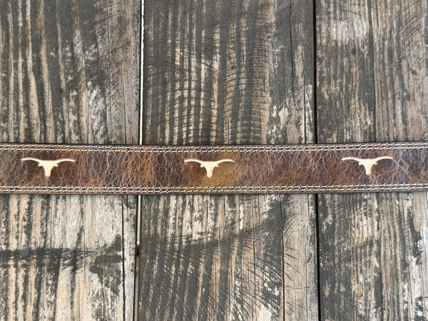 A close-up of a brown leather belt with three light-colored steer skull emblems, laid flat on a rustic weathered wood plank surface. Available at Blair's Western Wear, Marble Falls, TX