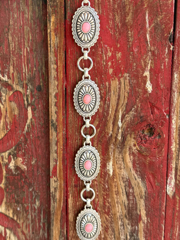 A1308230 — Kid's Pink Concho Chain Western Belt Full silver concho chain belt with repeating oval silver conchos featuring engraved petal/sunburst detail, rope edge border, and pink stone center in each. Silver O-ring links connect each concho. Adjustable silver O-ring chain tail with small pink stone drop tip. Available at Blair's Western Wear located in Marble Falls TX 