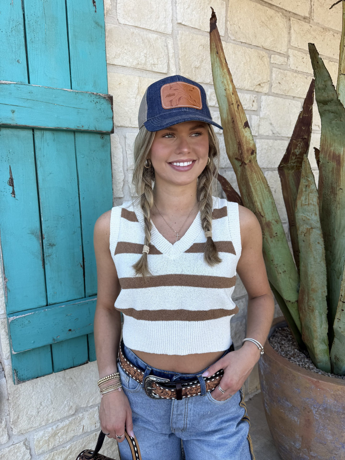 Woman wearing a striped sleeveless top and cap, standing next to a plant and blue door.