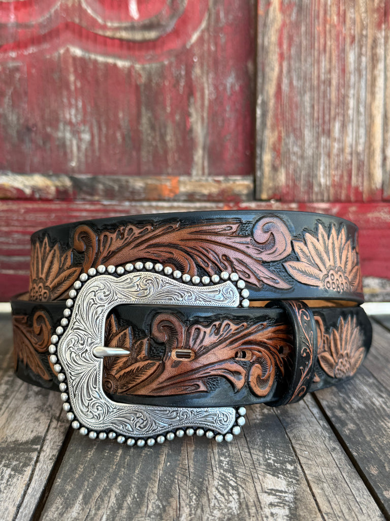 Black leather hand-tooled with cognac floral and sunflower scroll detailing throughout, finished with a large ornate silver scroll buckle bordered with silver studs. Bold contrast, artisan craftsmanship, and serious Western style — all in one belt. Shop at Blair's Western Wear — blairswesternwear.com.