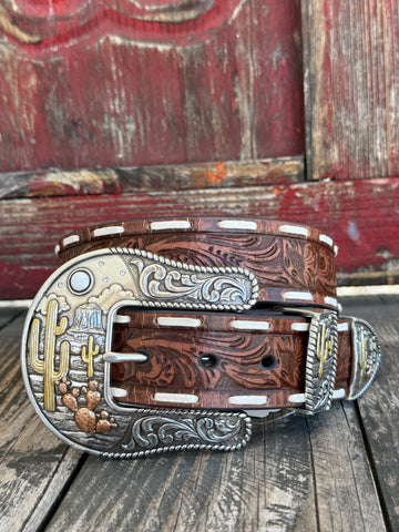 A western-style leather belt with a carved brown leather body, silver-toned engraved buckle, and white contrast stitching, displayed on a rustic wooden surface in front of a weathered red wooden door.