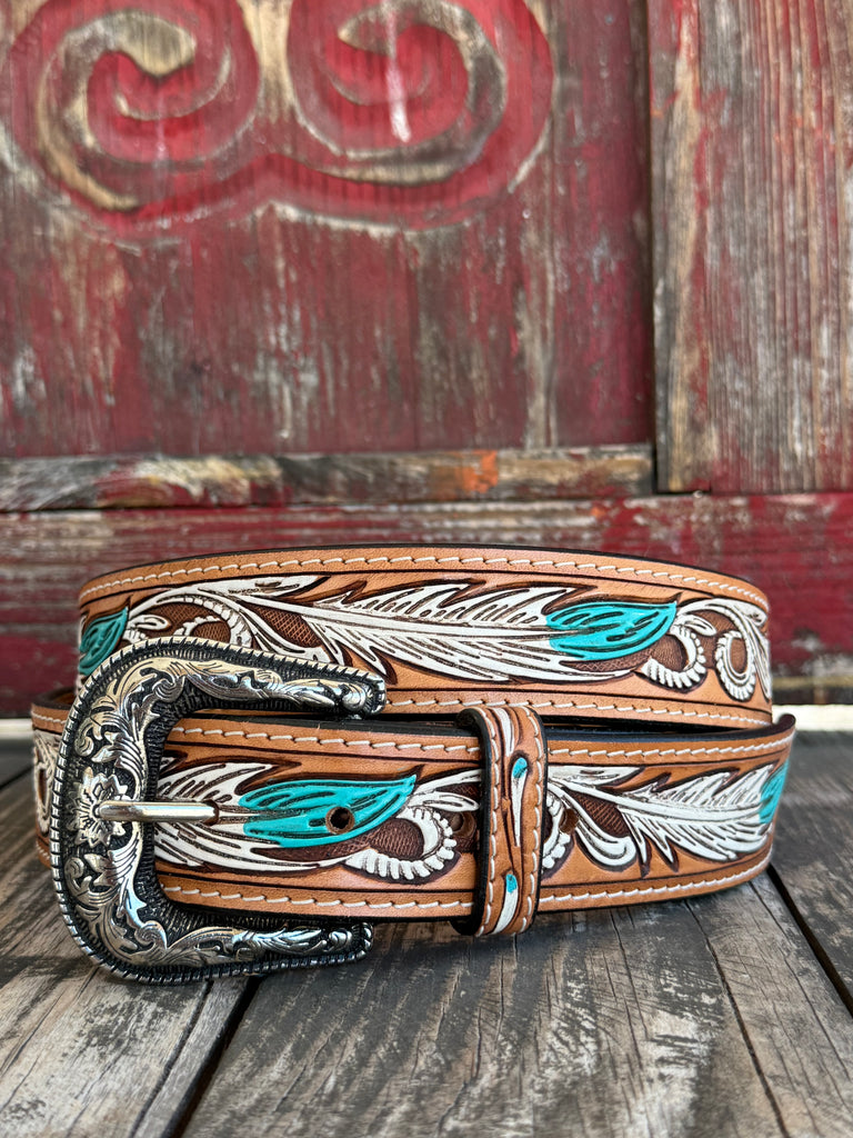 A tooled brown leather belt with turquoise and white feather designs, featuring an ornate silver buckle, displayed on a rustic wooden surface in front of a weathered red wooden door.