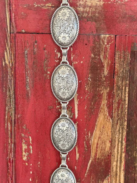 Linked oval silver conchos, each hand-engraved with flowing floral scroll detail and framed by a rope border, create a fully metal concho chain belt that makes a stunning western statement — no leather strap, just pure silver artistry from end to end. Available at BLAIR'S Western Wear located in Marble Falls TX 
