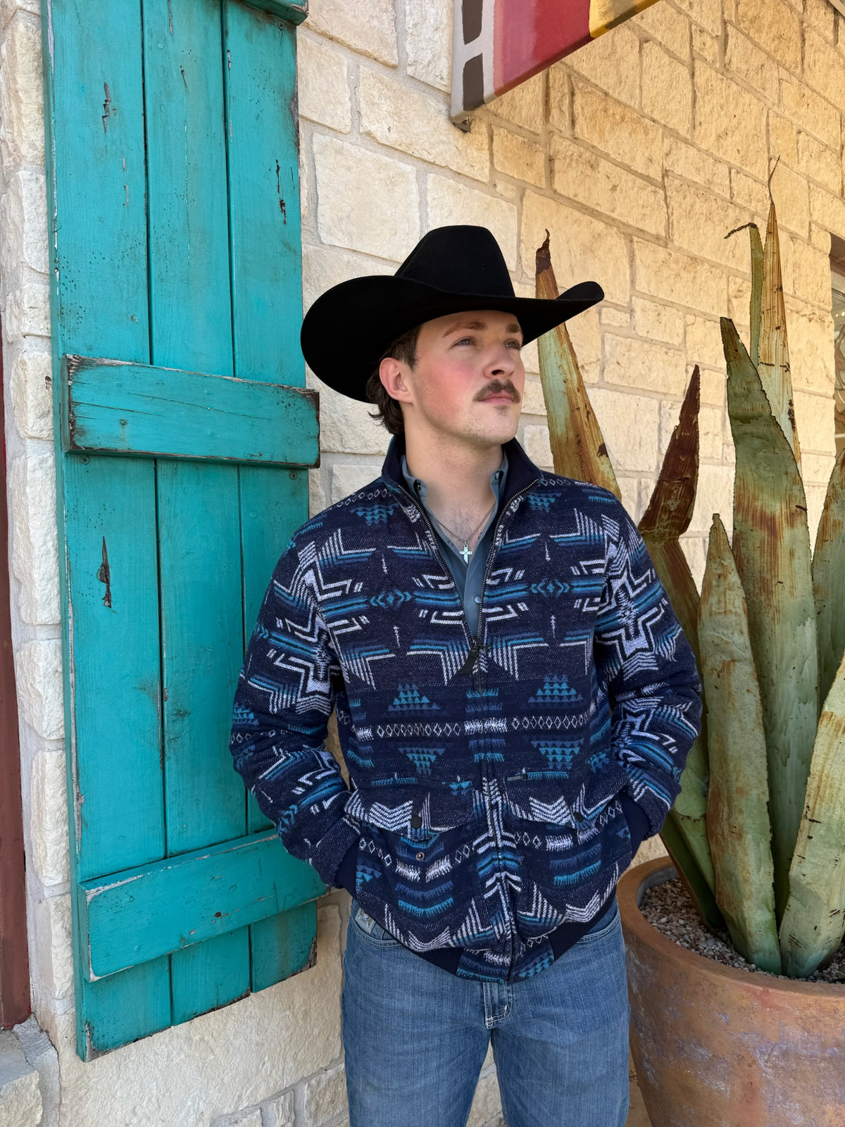 Man wearing a cowboy hat and patterned shirt standing in front of a building with turquoise shutters and a cactus plant.