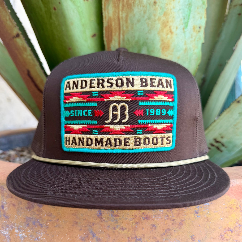 A dark brown snapback cap with a large embroidered front patch reading 'ANDERSON BEAN' and 'HANDMADE BOOTS,' photographed outdoors against green plant leaves.