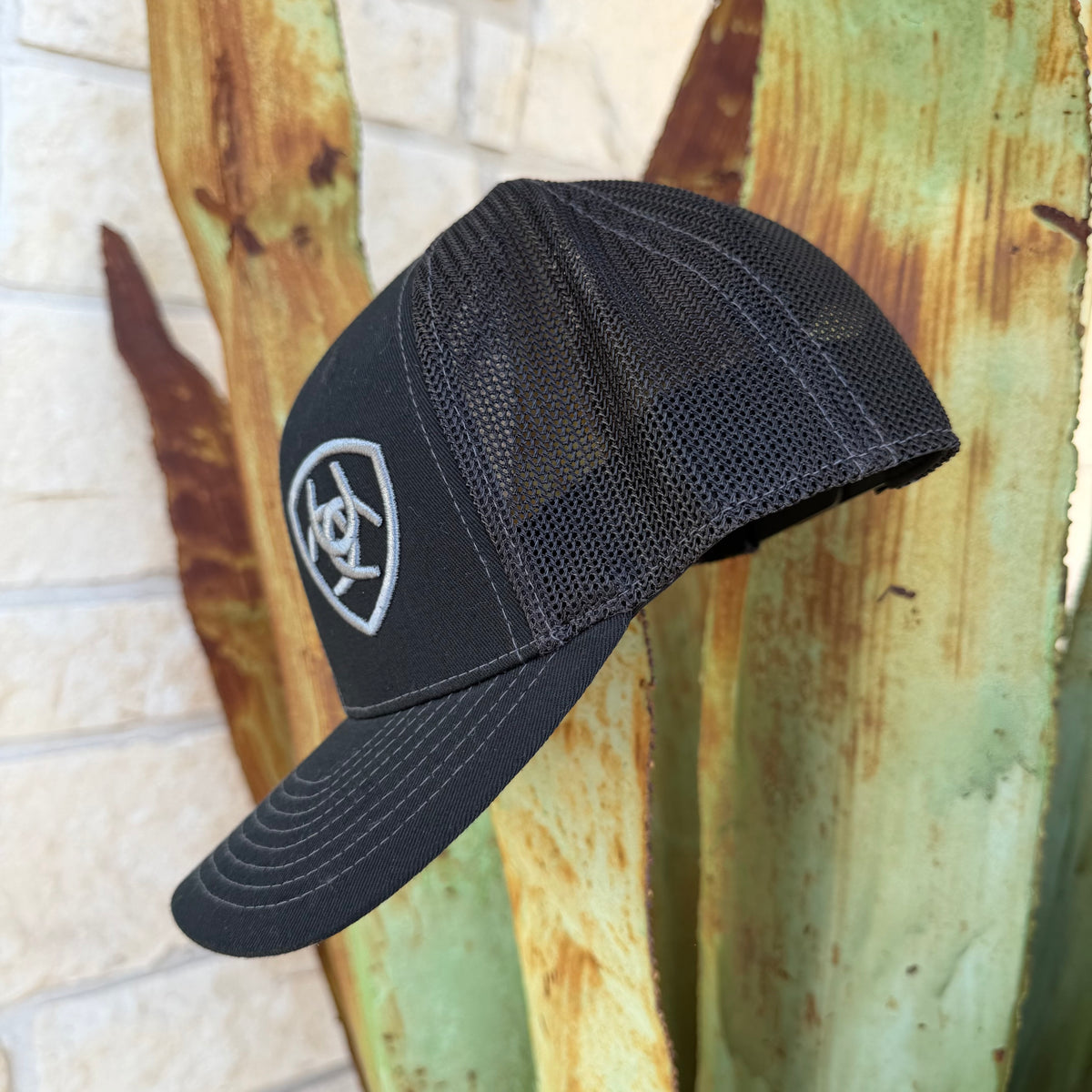 A black trucker-style baseball cap with a mesh back and a gray shield logo on the front,