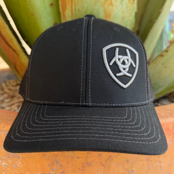 A black trucker-style cap with a silver embroidered shield logo on the front, 
