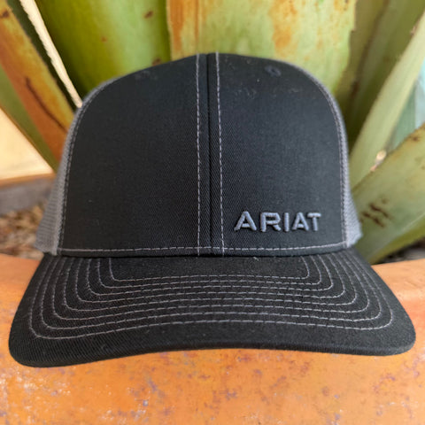 A black trucker-style cap with gray mesh side panels and gray contrast stitching, featuring the word 'ARIAT' embroidered on the front. 