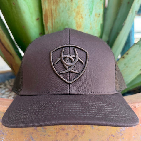 A dark brown trucker hat with a matching embroidered shield logo on the front, displayed outdoors against a green agave plant and a rusted orange surface.
