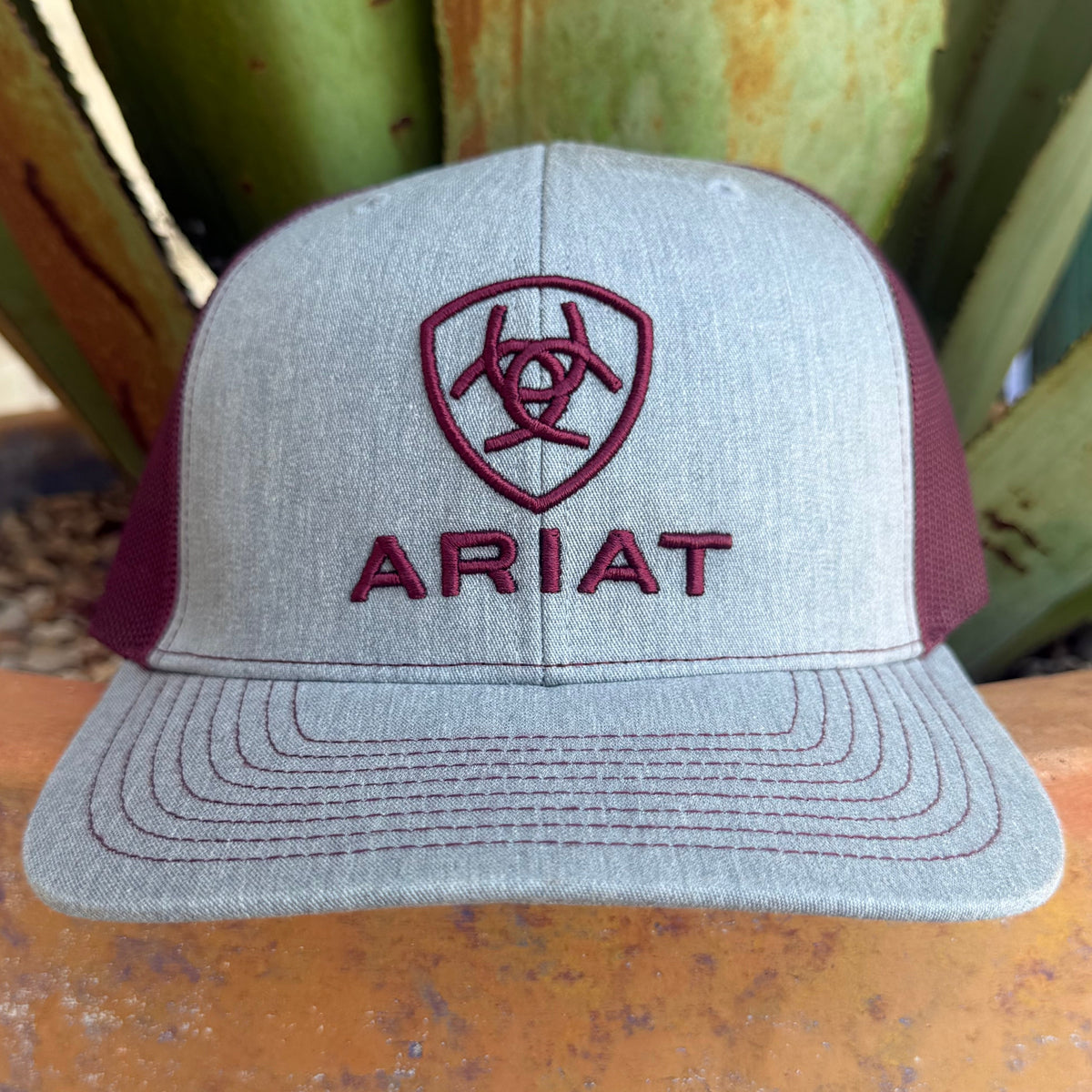 A light gray heather trucker hat with maroon mesh side panels and matching maroon embroidery featuring the Ariat   logo and the word ARIAT,