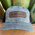 A heather gray trucker hat with black mesh side panels and a brown leather patch on the front that reads ARIAT, 