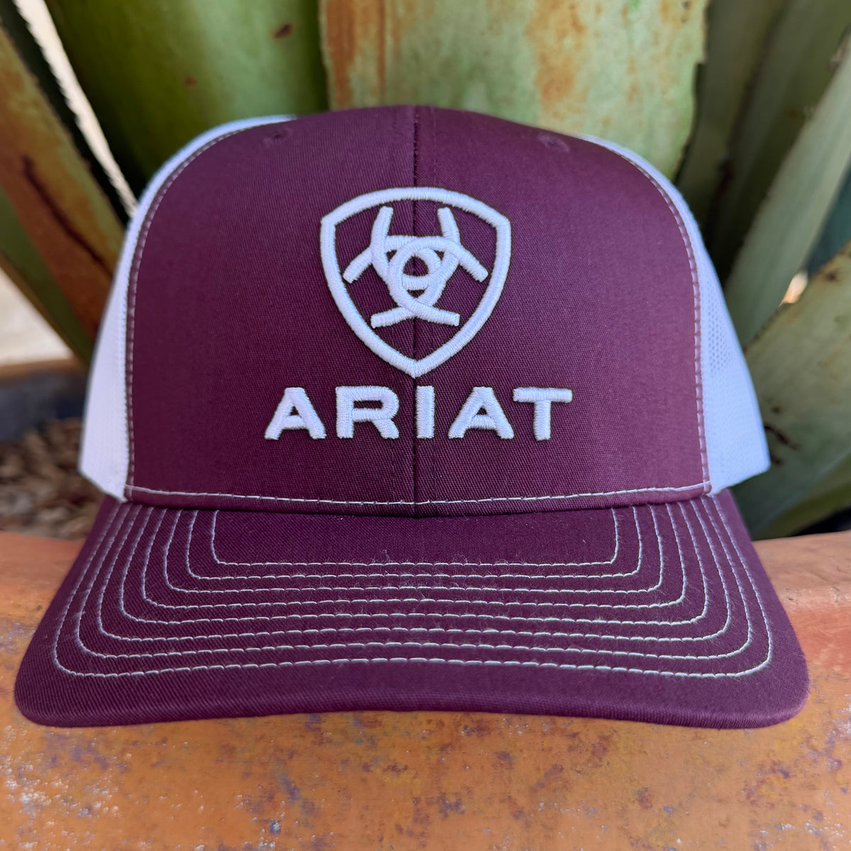 A maroon and white trucker hat with white embroidered Ariat logo and text, 
