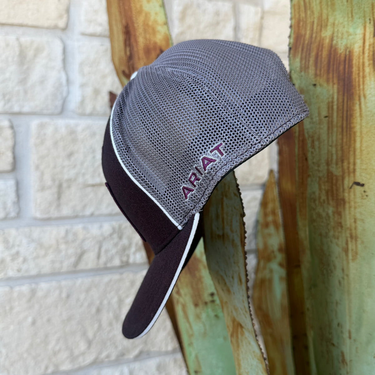 A gray and dark burgundy trucker-style cap with a mesh back and a dark visor, The word 'ARIAT' is embroidered in burgundy on the side 
