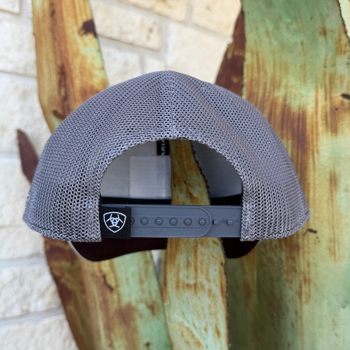 A gray mesh-back trucker cap shown from the back, featuring an adjustable dark gray strap with a black patch displaying the ARIAT logo