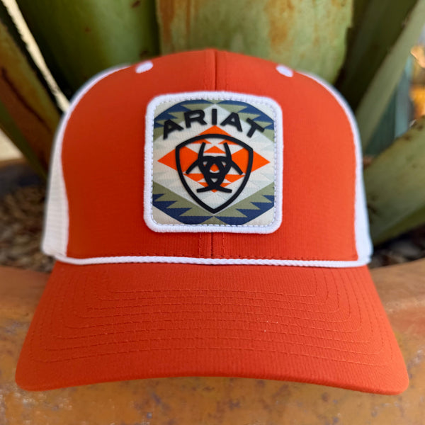 An orange and white trucker hat with a white patch on the front that reads 'ARIAT' and features an aztec inspired pattern 