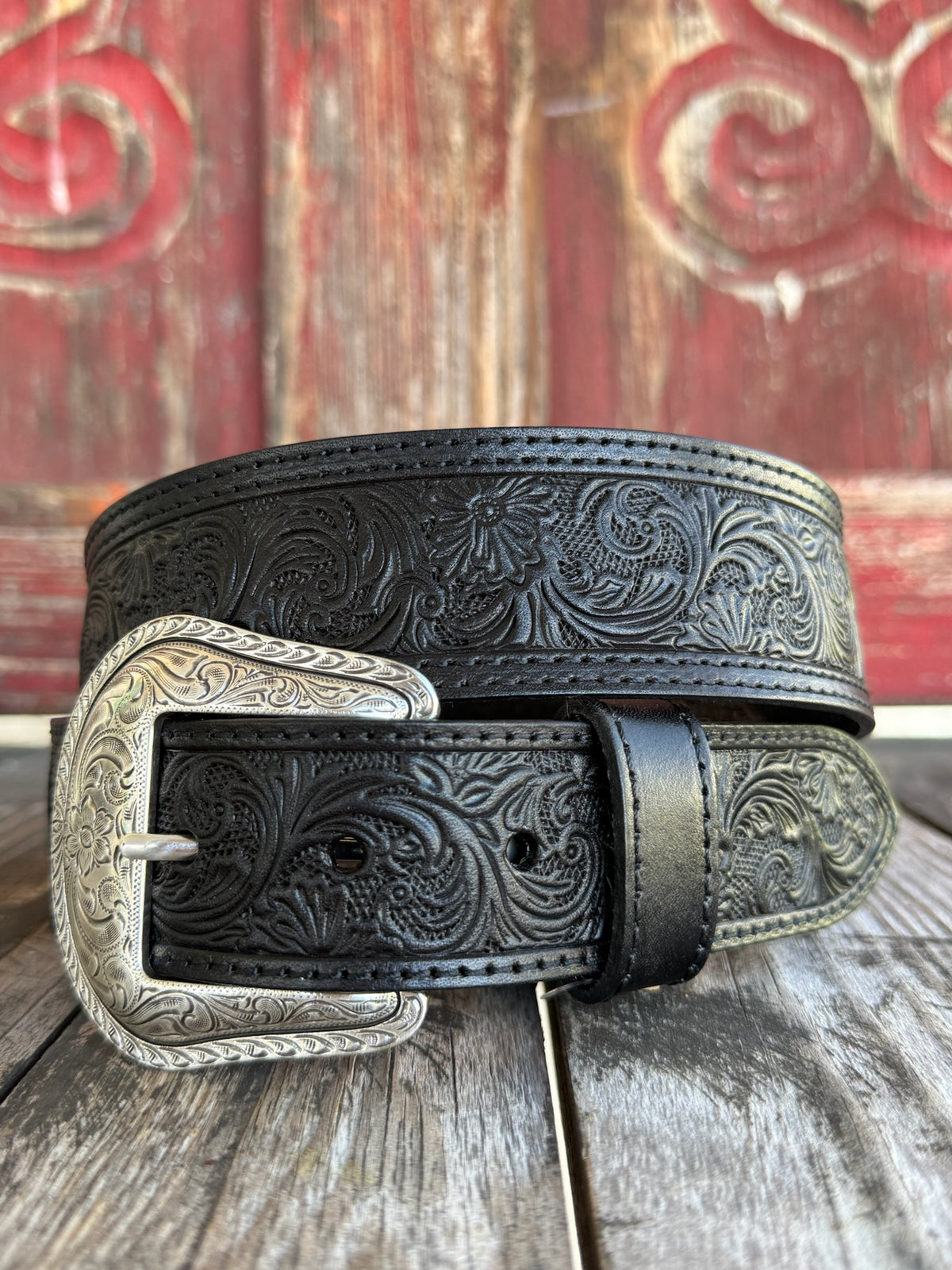 Black leather with edge-to-edge floral scroll tooling, dot edge stitching, and a large engraved silver western buckle with matching keeper — a timeless western belt with deep, handcrafted detail and sharp black-on-silver contrast.