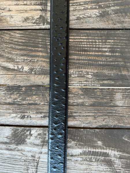A black ostrich leather belt with a large, ornate silver-toned western buckle, coiled on a rustic wooden surface with a weathered red wooden background.