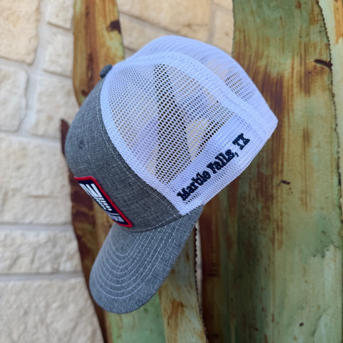 A gray and white trucker-style cap with a white mesh back, embroidered 'Marble Falls, TX' text on the side, and a red-and-black patch on the front, 